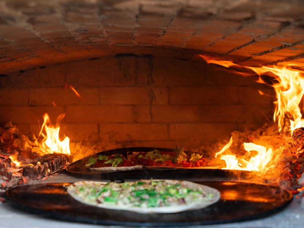 Blick in den Holzbackofen. Es ist ein frisch belegter Flammenkuchen zu sehen und an den Seiten sind noch Flammen sichtbar.