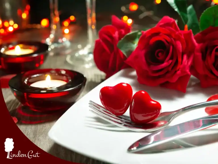Liebevoll gestalteter Tisch mit einem weißen, eckigen Teller auf dem Besteck liegt.. Auf der Gabel befinden sich zwei rote Herzen. Es sind Teelichter in roter Halterung und rote Rosen zu sehen.