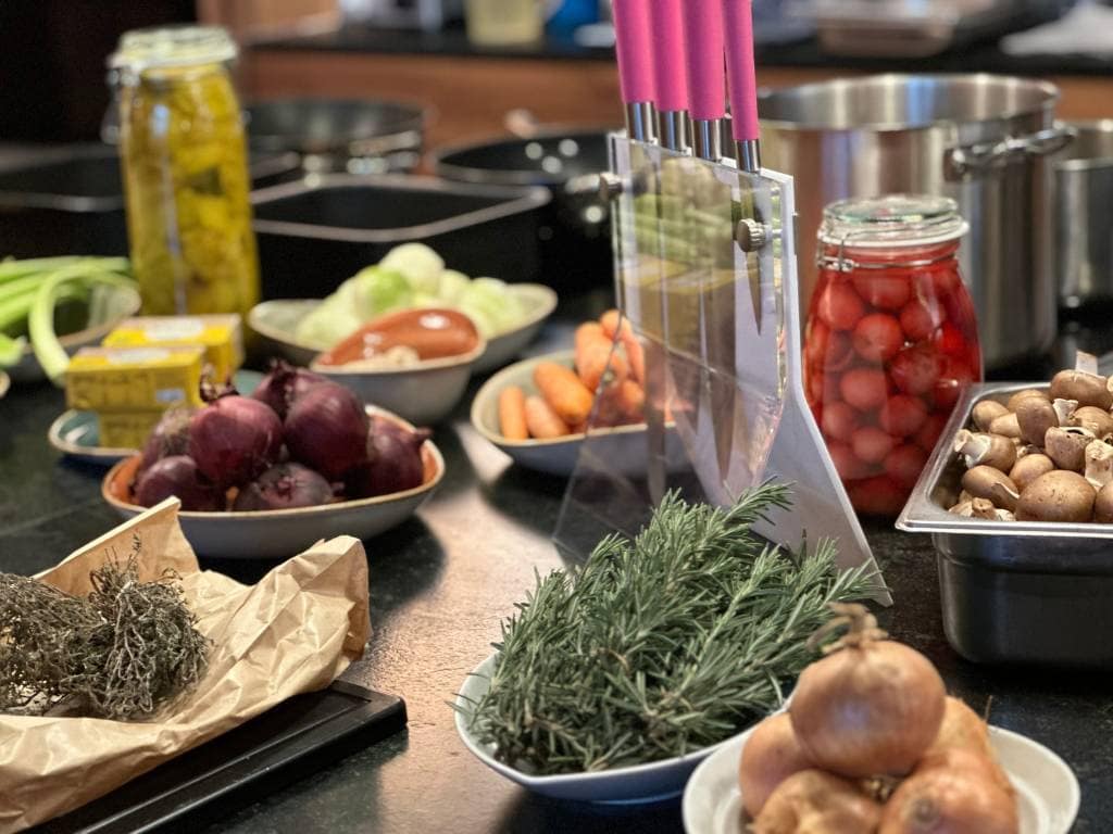 Aufgebaute Zutaten für für den Kochkurs mit Zwiebeln, Kräutern, eingelegten Radieschen, einem Messerblock und im Hintergrund ist Kochgeschirr zu sehen.